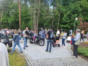 Dwoje dzieci siedzących na motocyklu a obok grupa ludzi zgromadzonych na tle drzew